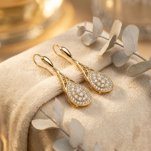 Gold-plated cubic zirconia drop earrings on soft cream fluff beside pale eucalyptus sprigs