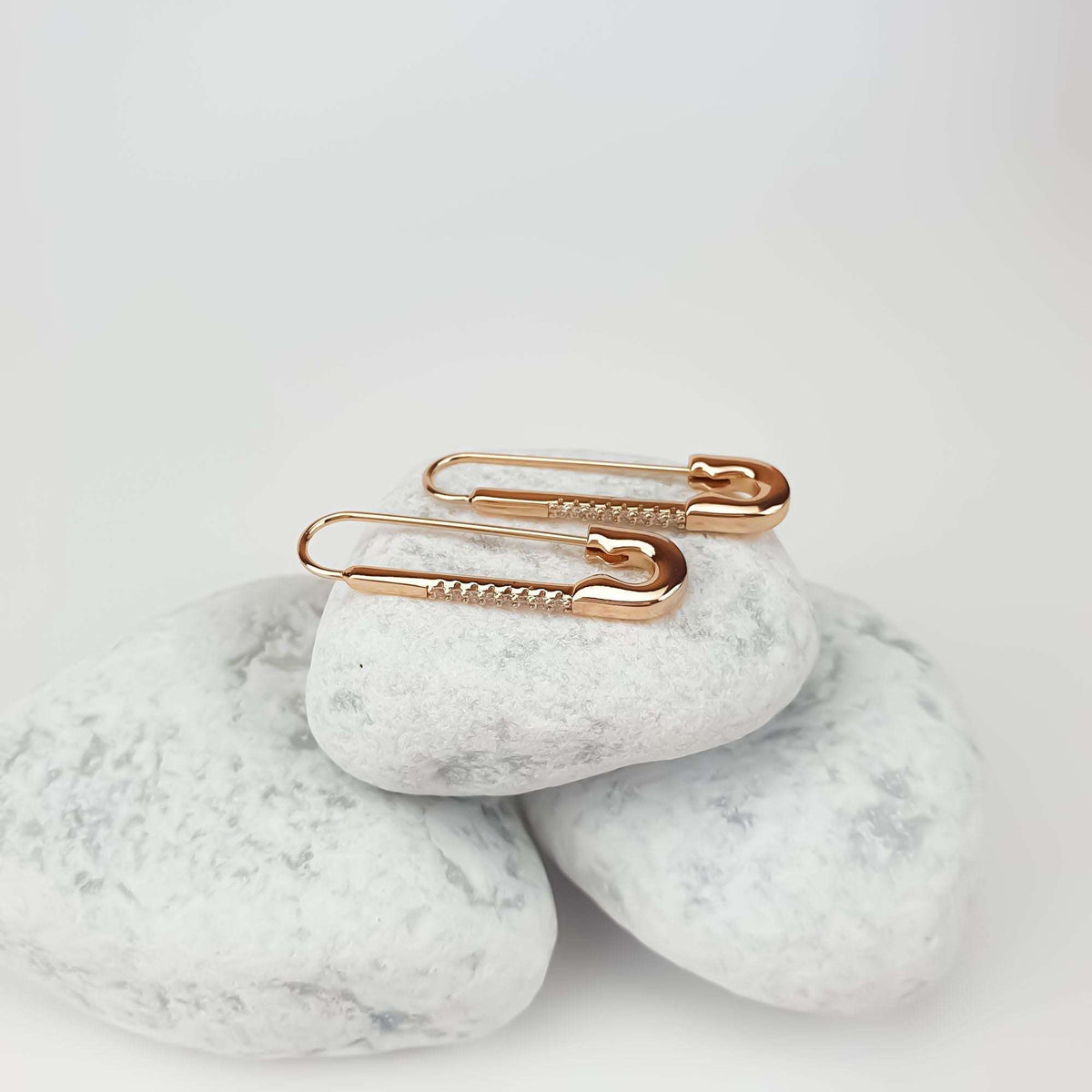 Pair of gold-tone safety pin earrings on white pebbles, clasp visible