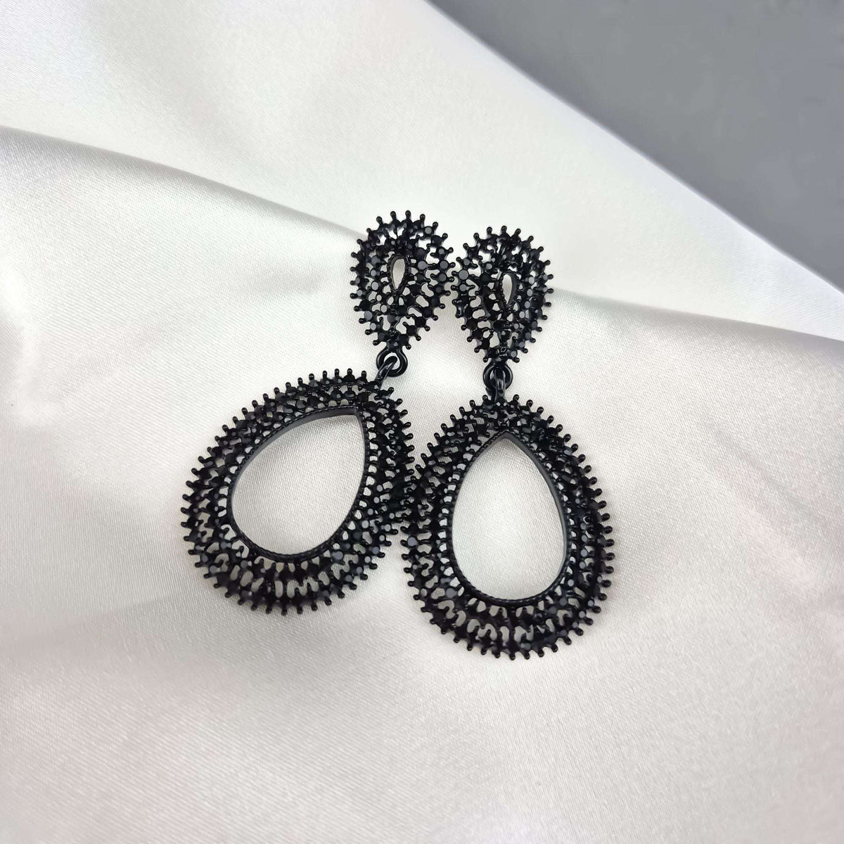 Two-part black openwork teardrop earrings at an angle on white silk drapery