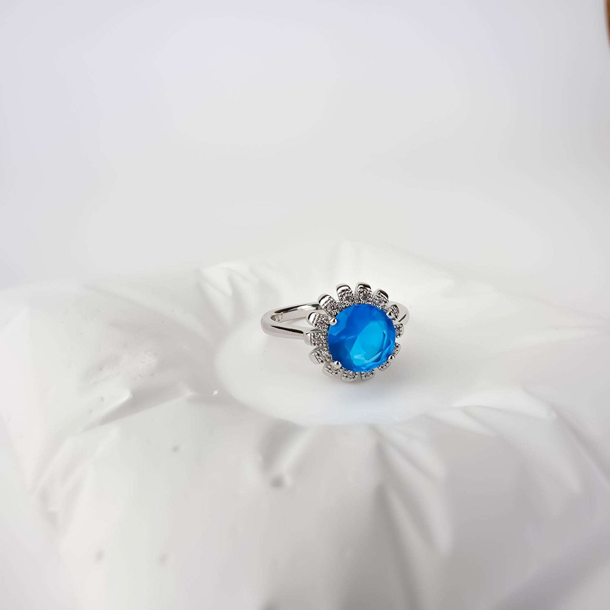 Platinum-tone ring with a round blue crystal in a flower setting on white fabric