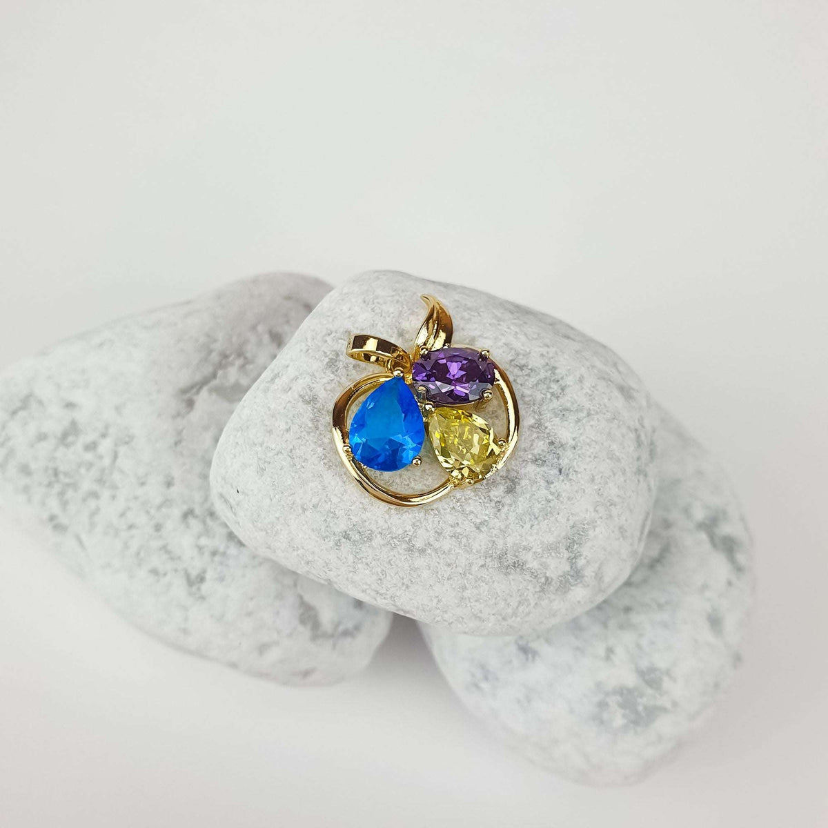 Gold-tone apple pendant with three bright stones at an angle on pale pebbles