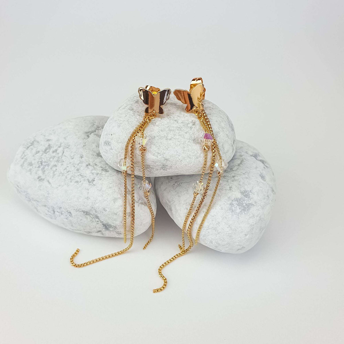 Gold-plated butterfly earrings with clear crystals and long chains on white pebbles