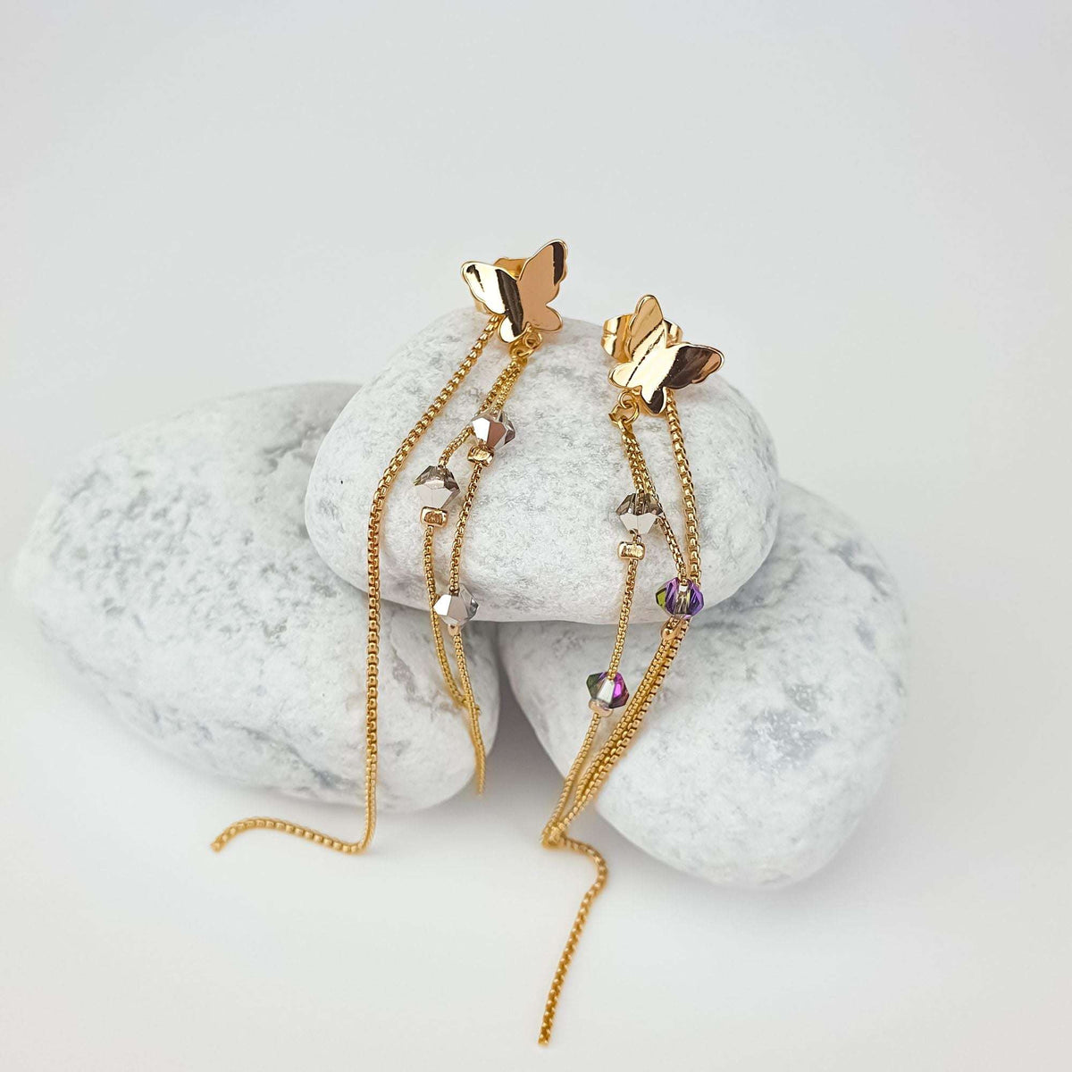 Gold-plated butterfly earrings with smoky grey crystals on fine chains, on white pebbles