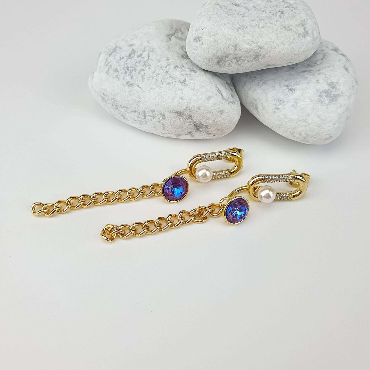 Earrings laid out flat, gold-plated chains with violet-blue crystals, on a pale surface
