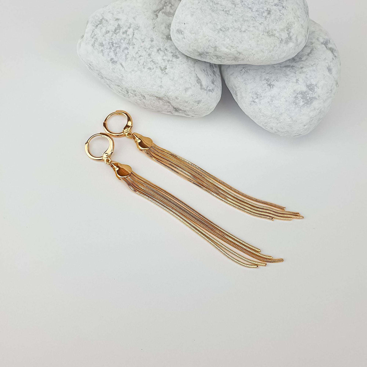 Gold-plated fringe earrings laid out beside white pebbles, their full length visible