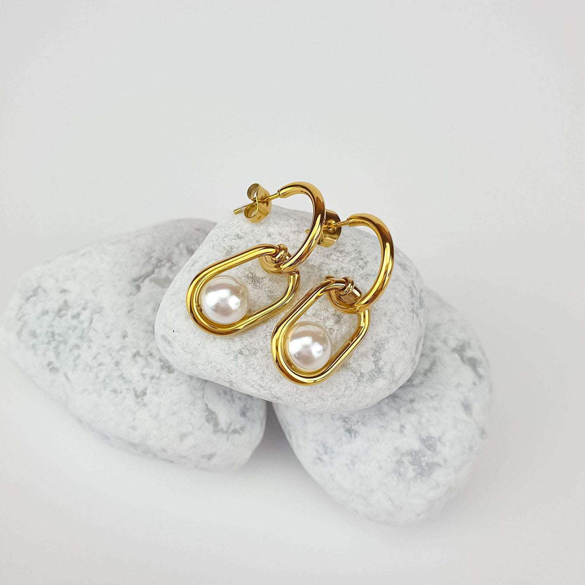 Curved gold-plated earrings with a faux pearl in an oval link, on white pebbles