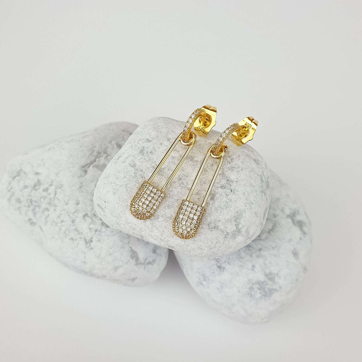 Gold-plated safety pin earrings with cubic zirconia and push-back studs on white pebbles