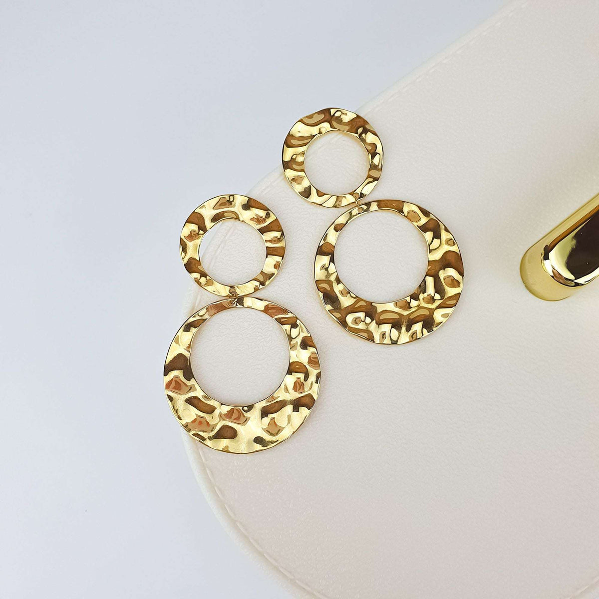 Gold-plated double-circle earrings with a hammered texture, angled on a cream box