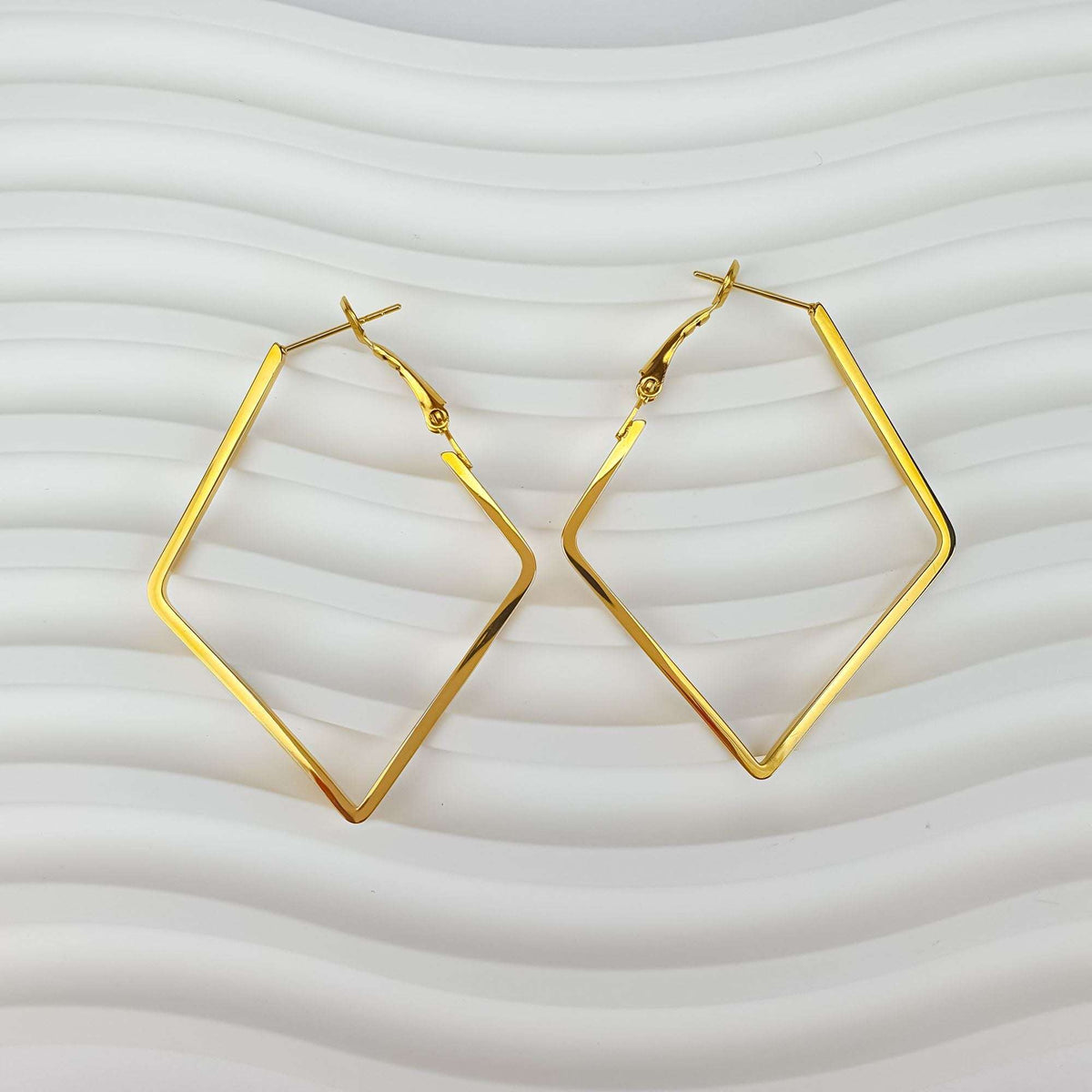 Gold-plated rhombus earrings with visible clasp on a wavy white surface