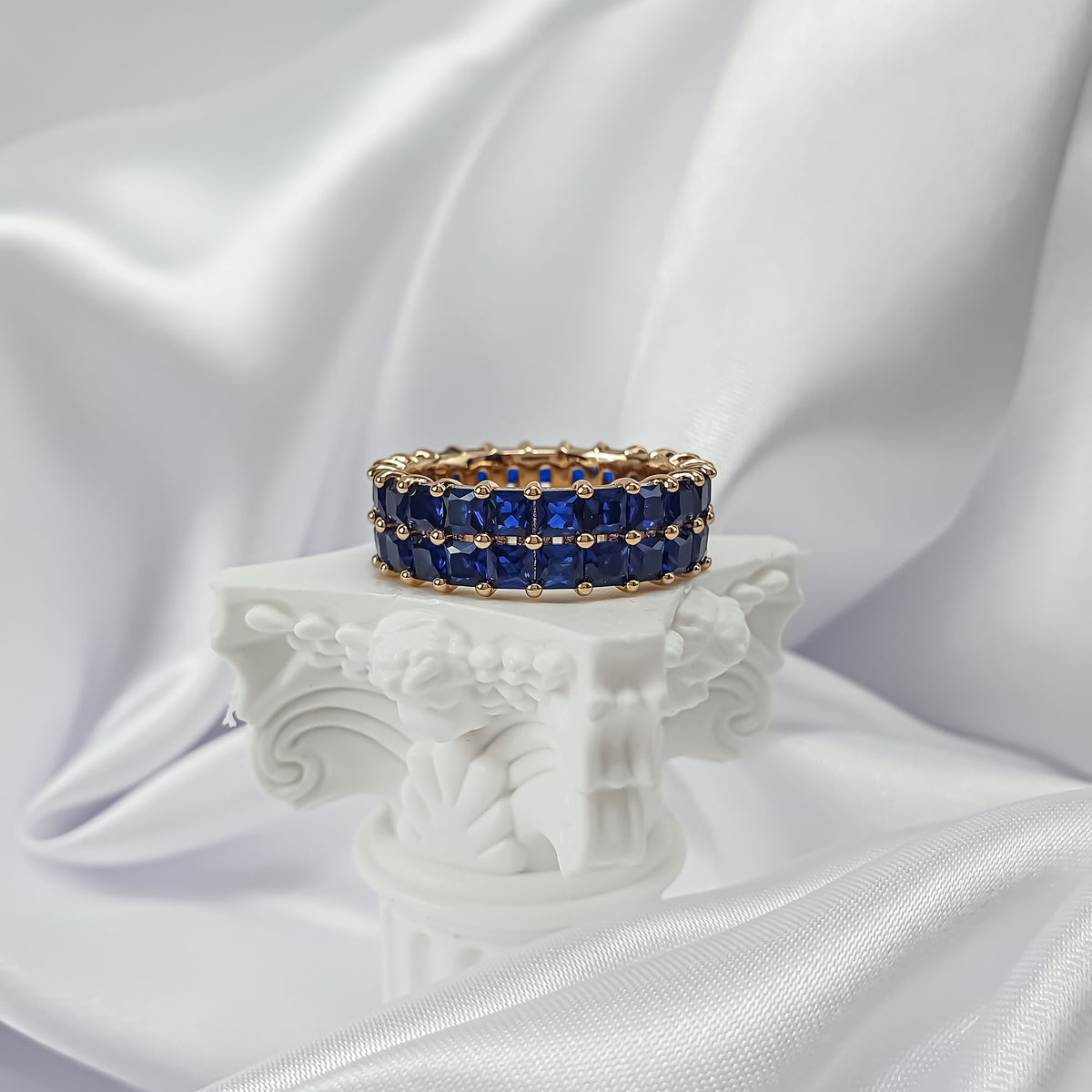 Dark blue crystal ring with gold plating on a white pedestal beside silky fabric