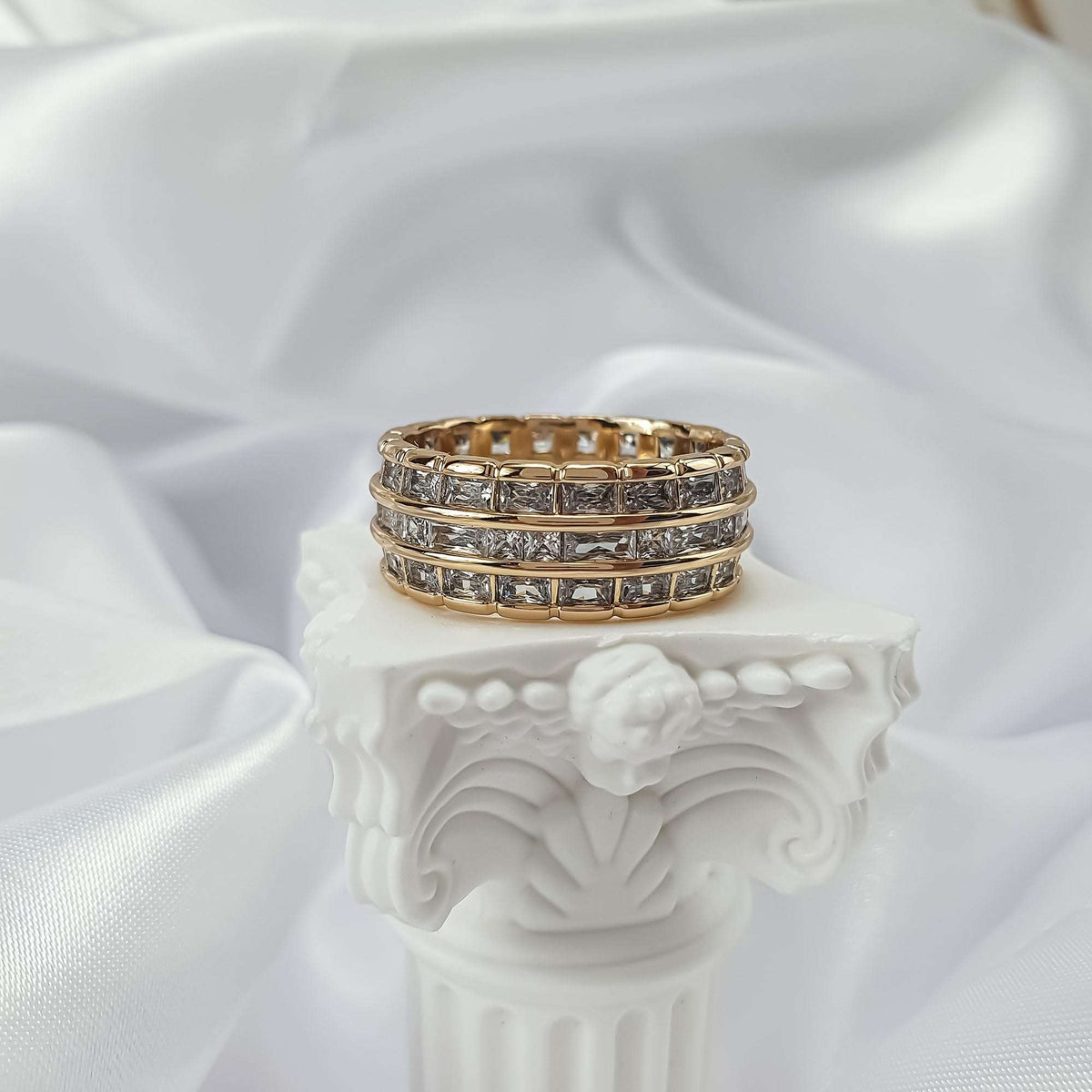 Gold-tone ring with three rows of clear stones on a white column, silk behind