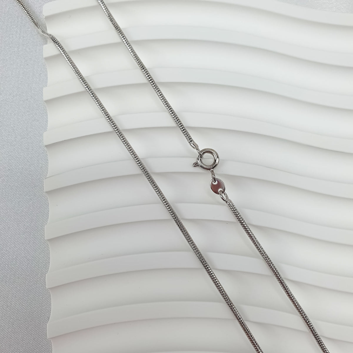 Smooth silver-tone snake chain laid on a white wavy surface, close up