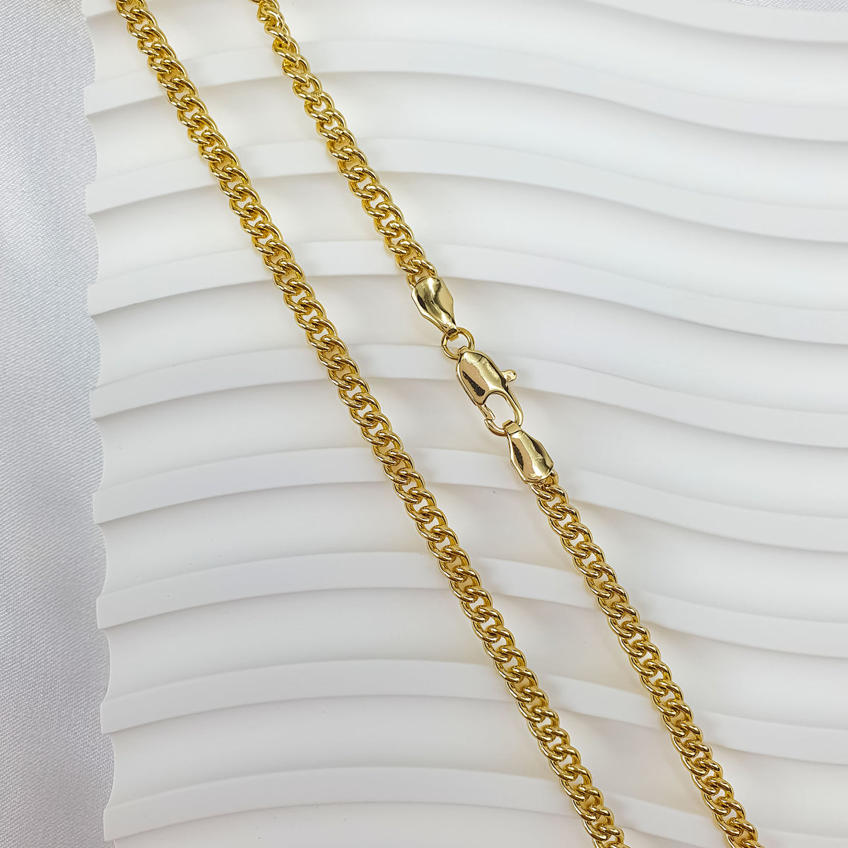 Polished gold-tone curb chain on a white wavy background with the clasp visible