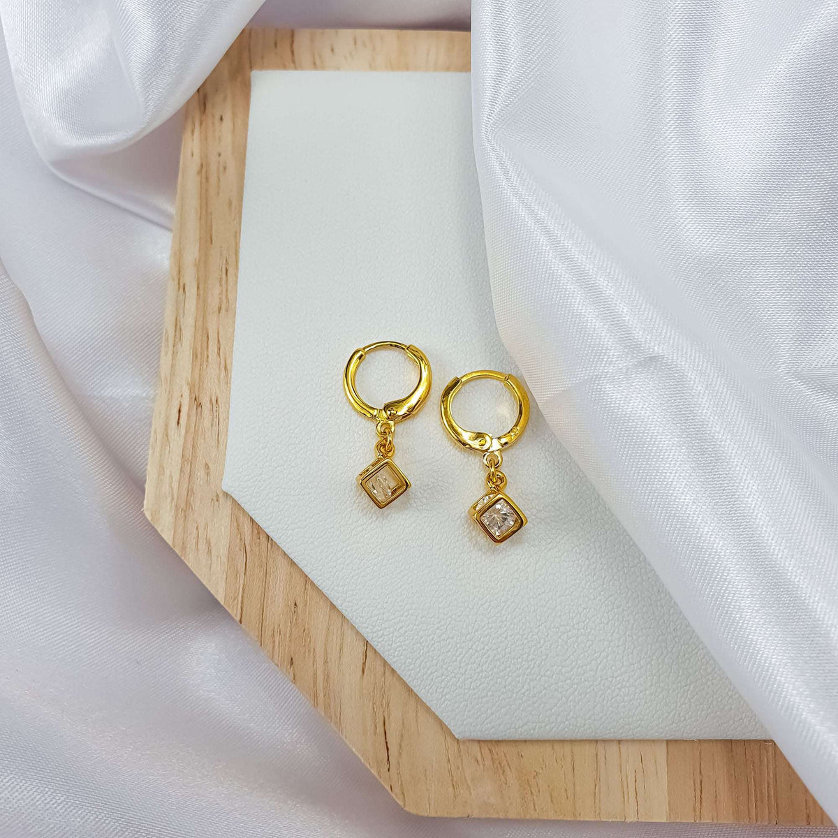 Pair of small gold-plated hoops with square crystal charms on a wooden stand