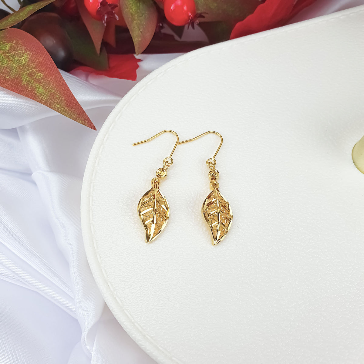 Gold-plated leaf earrings with fish hooks on a white stand with berries
