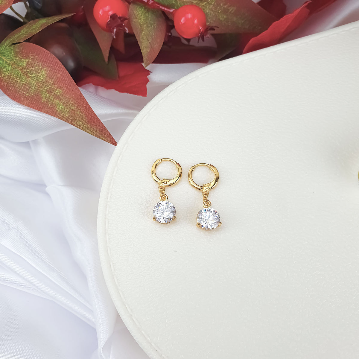 Gold-plated earrings with bright stones on a white stand among red berries