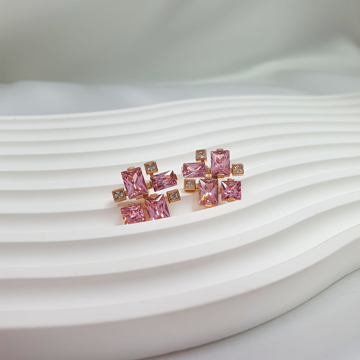 Pink cubic zirconia stud earrings with a gold-tone frame on a wavy white surface