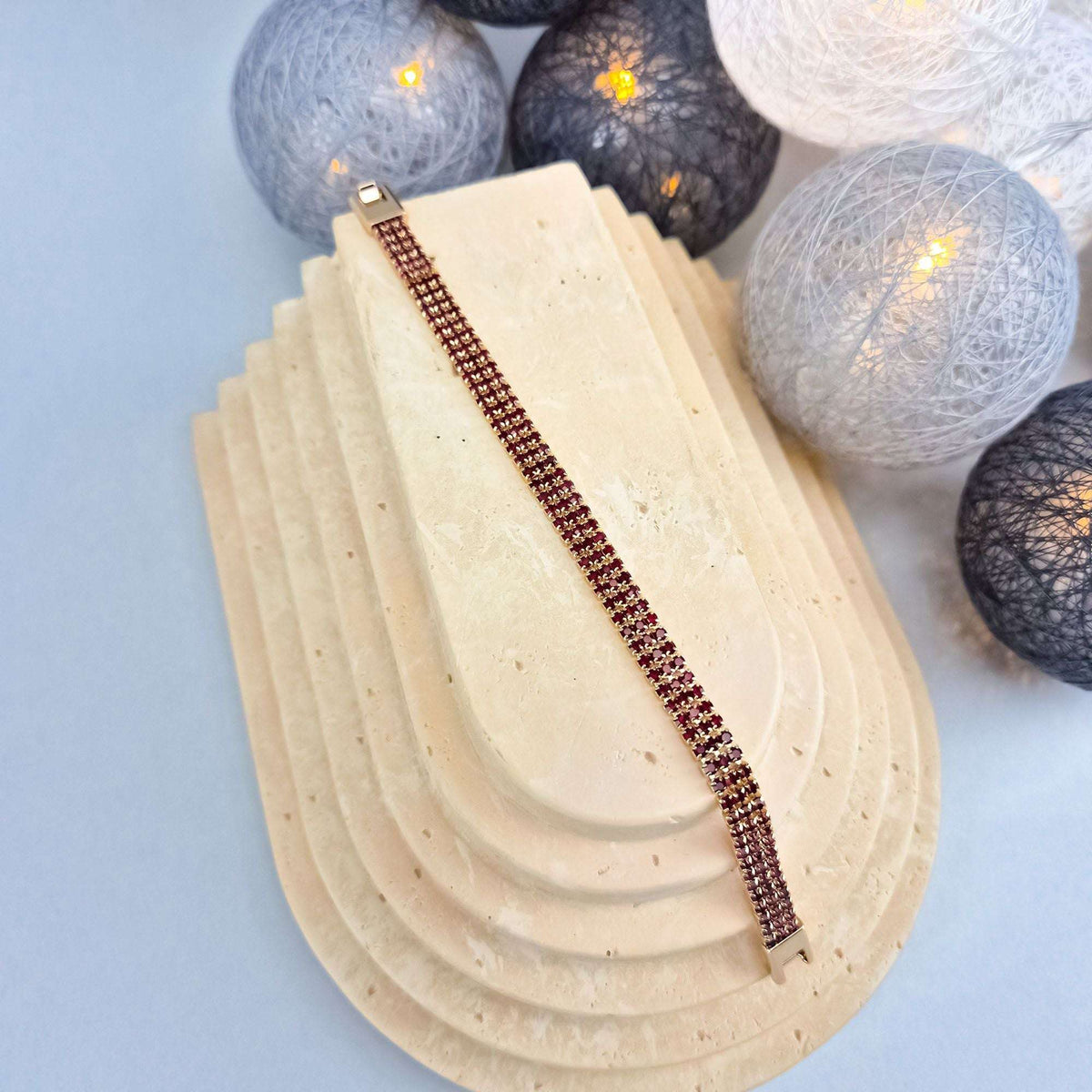 Gold-plated red cubic zirconia bracelet laid out on a sand-coloured stand, warm lights behind