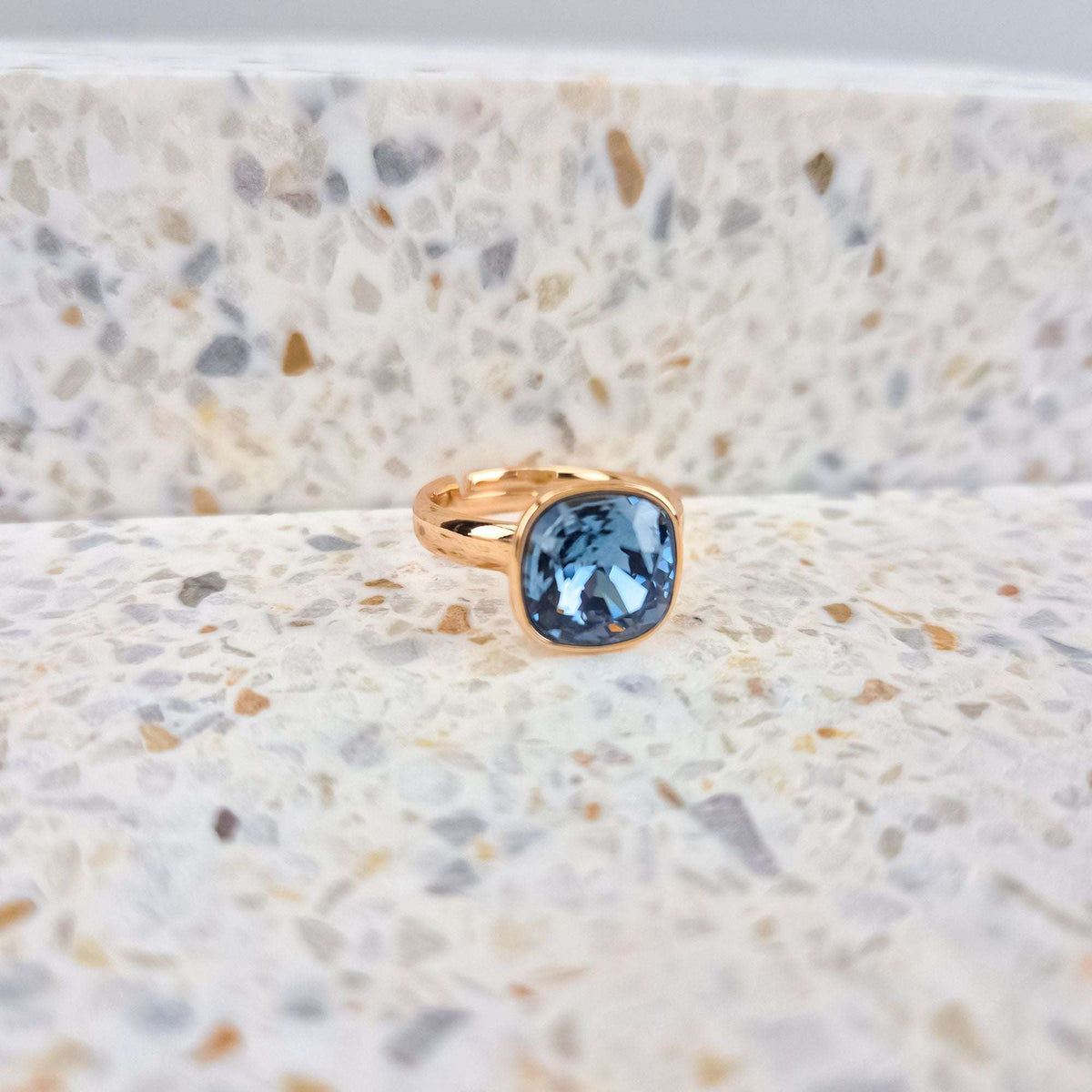 Gold-plated ring with a cushion-shaped blue crystal, close-up on speckled stone