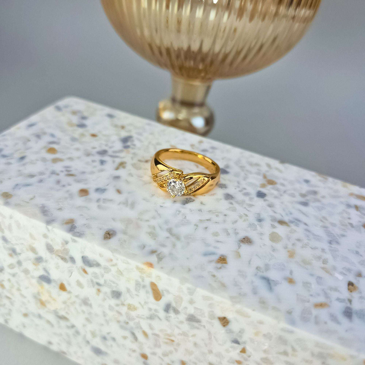 Gold-tone cubic zirconia ring on terrazzo with a ribbed vase behind