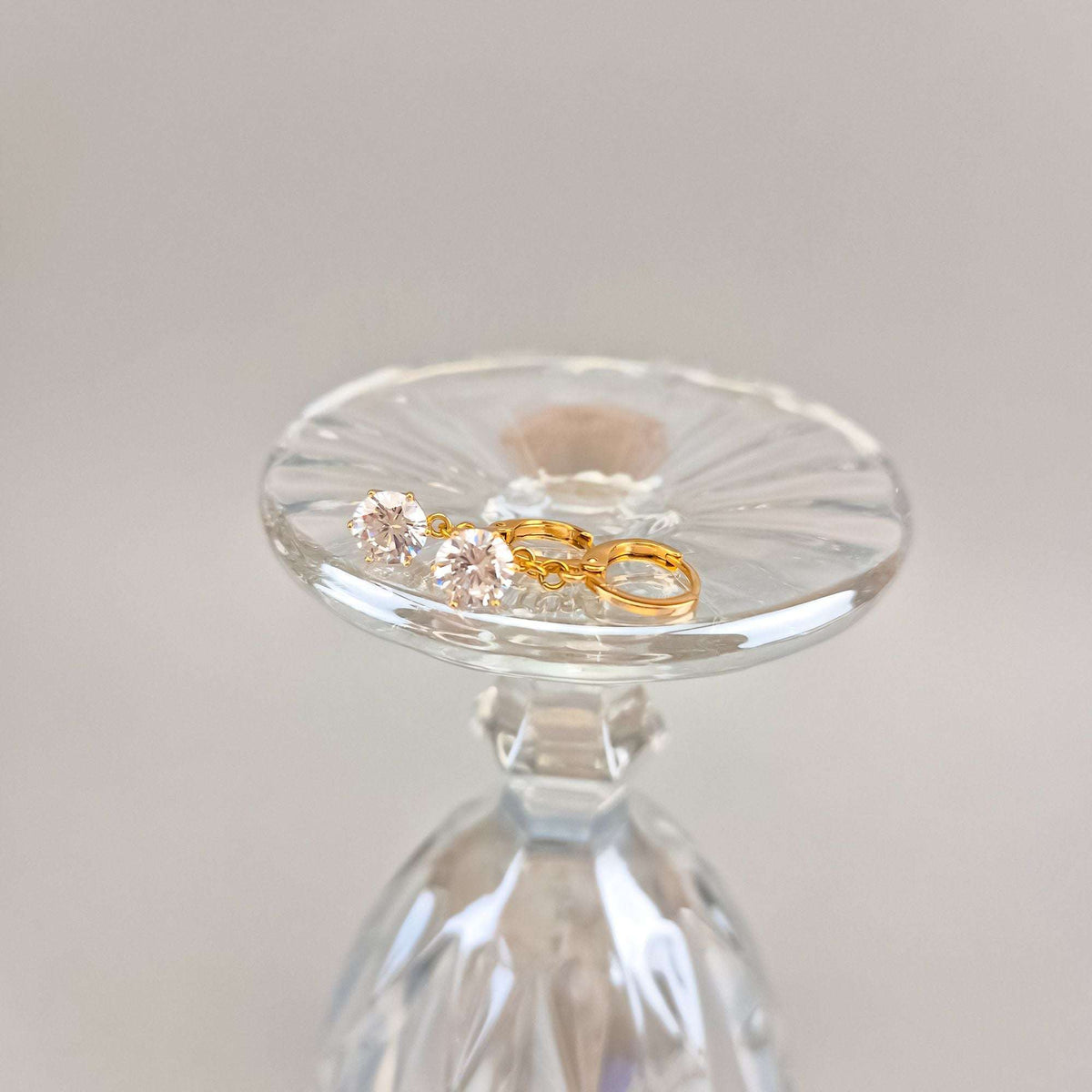 Gold-plated earrings with round clear stones on a glass stand, soft beige background