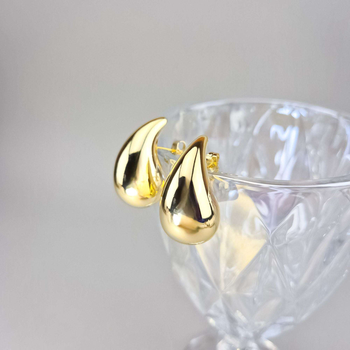 Puffy gold-plated teardrop earrings resting on the rim of a crystal glass