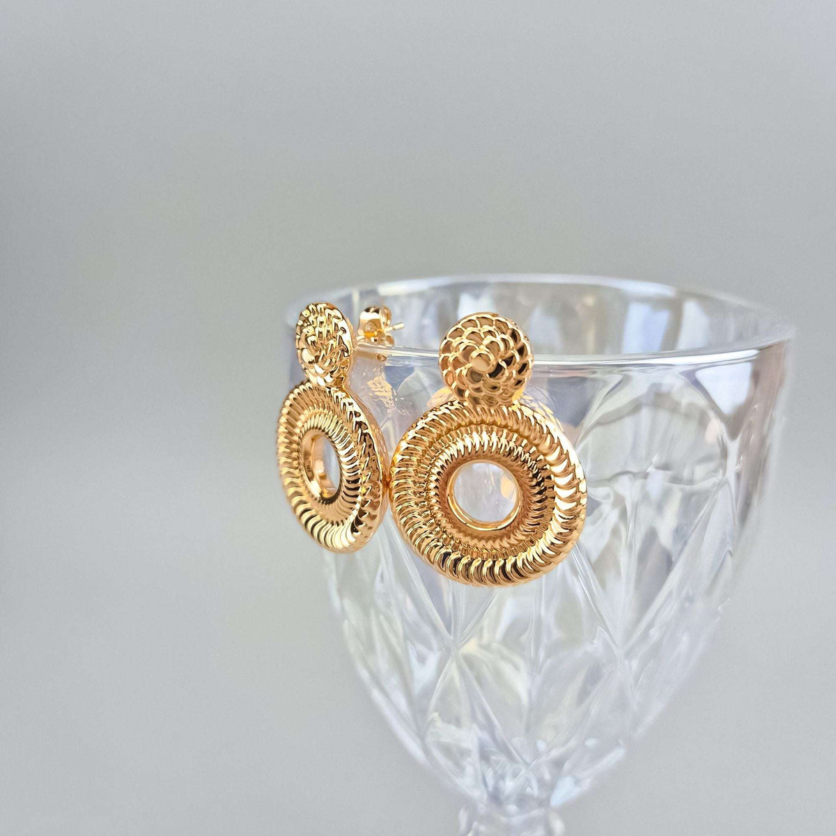 Two gold-plated round earrings with push-back studs on the rim of a glass