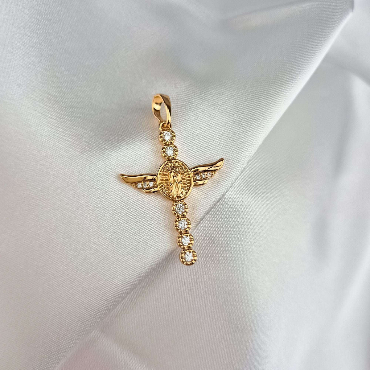 Winged cross pendant with cubic zirconia in a warm gold tone on white satin